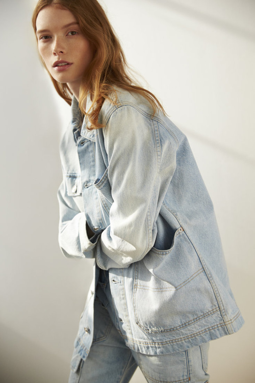 A red-haired female model wearing a light blue denim jacket and jeans poses against a neutral wall indoors.