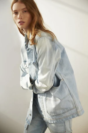 A red-haired female model wearing a light blue denim jacket and jeans poses against a neutral wall indoors.