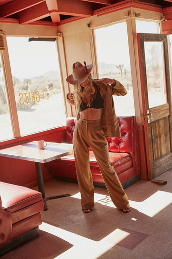 A woman in a brown suit and cowboy hat poses inside a vintage diner with desert scenery visible through the windows.