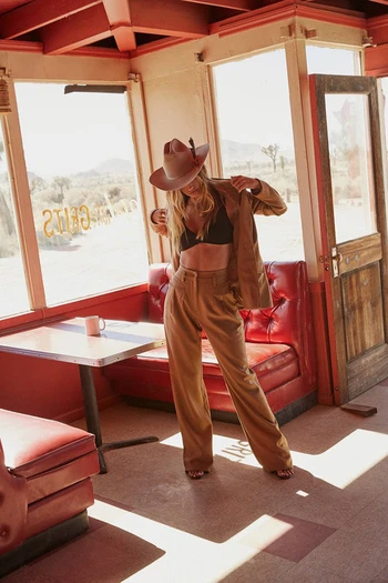 A woman in a brown suit and cowboy hat poses inside a vintage diner with desert scenery visible through the windows.