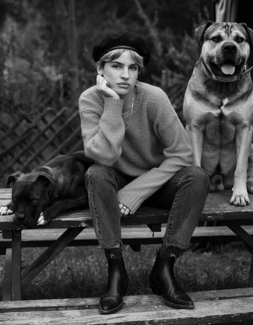 Black and white portrait of a woman in a beret sitting outside with two dogs on a wooden bench in a natural outdoor setting.
