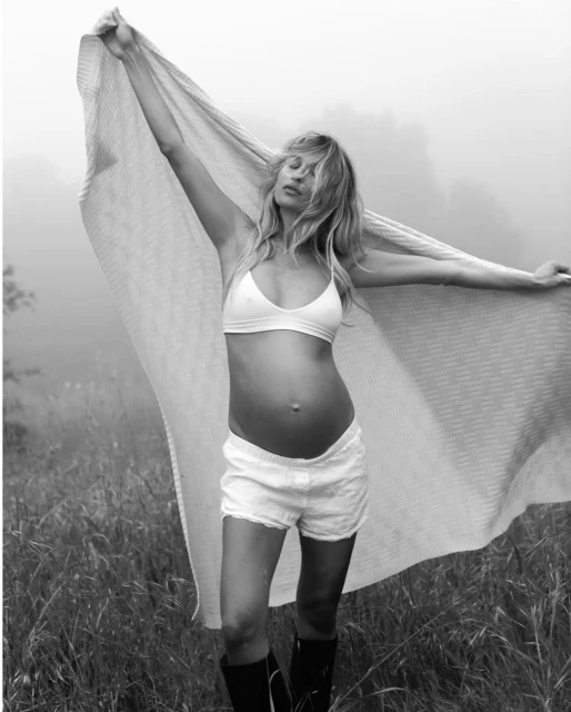 Black and white portrait of a pregnant woman standing in a field holding a blanket, wearing a bra top, shorts, and boots.