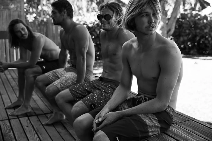 Four shirtless young men in boardshorts seated on a wooden outdoor deck in a black and white photograph by Sylve Colless.