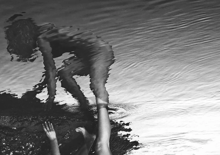 Black and white photo of a person standing in water near the shore, reflection distorted by water ripples in black and white.