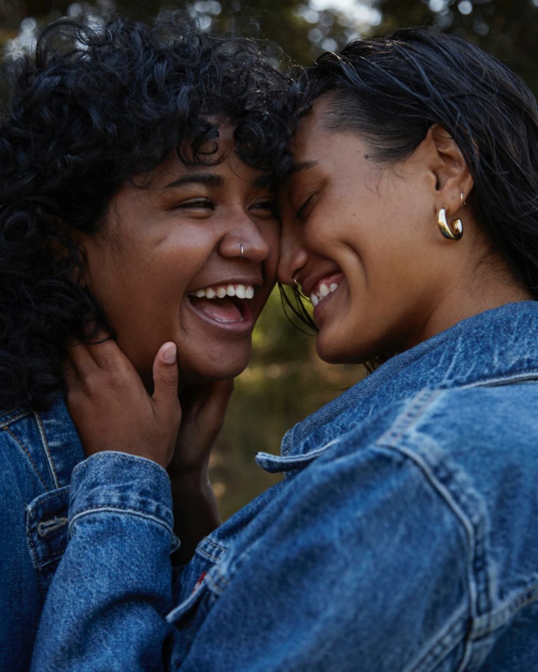 Two young women smile intimately outdoors, wearing denim jackets. One has an open mouth and curly hair, the other closed eyes.