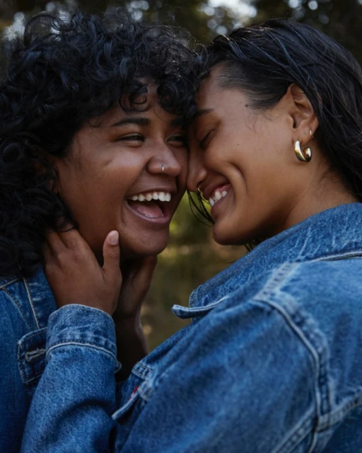 Two young women smile intimately outdoors, wearing denim jackets. One has an open mouth and curly hair, the other closed eyes.