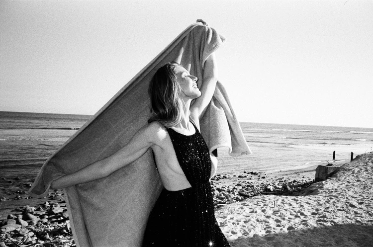 Woman in sequin dress stands on sandy beach, holding a towel overhead, ocean and sky visible in background, black and white photo.