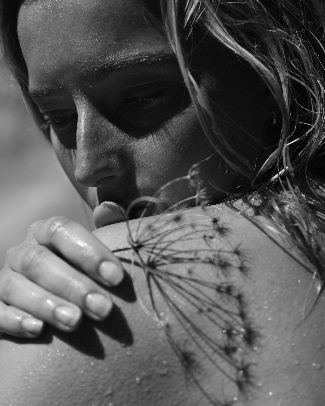 Monochromatic portrait of a young woman with wet hair, head turned, holding a flower against her back, shallow depth of field.