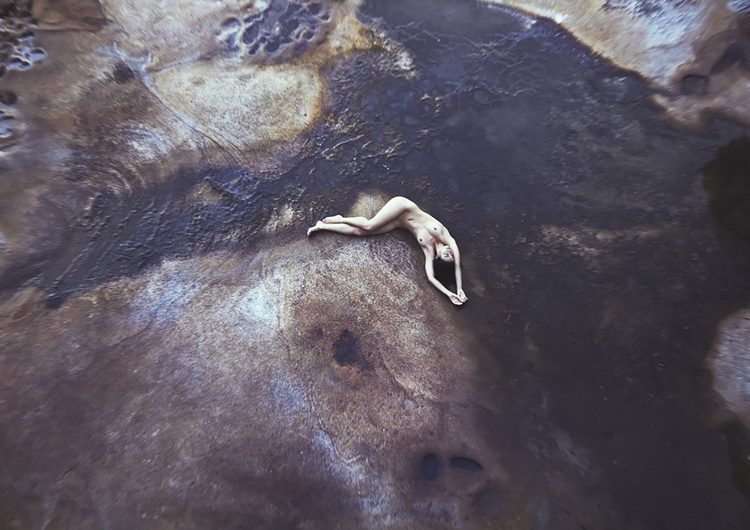 Aerial perspective of a nude adult female lying prone with arms raised on dark patterned textured rock formation outdoors.