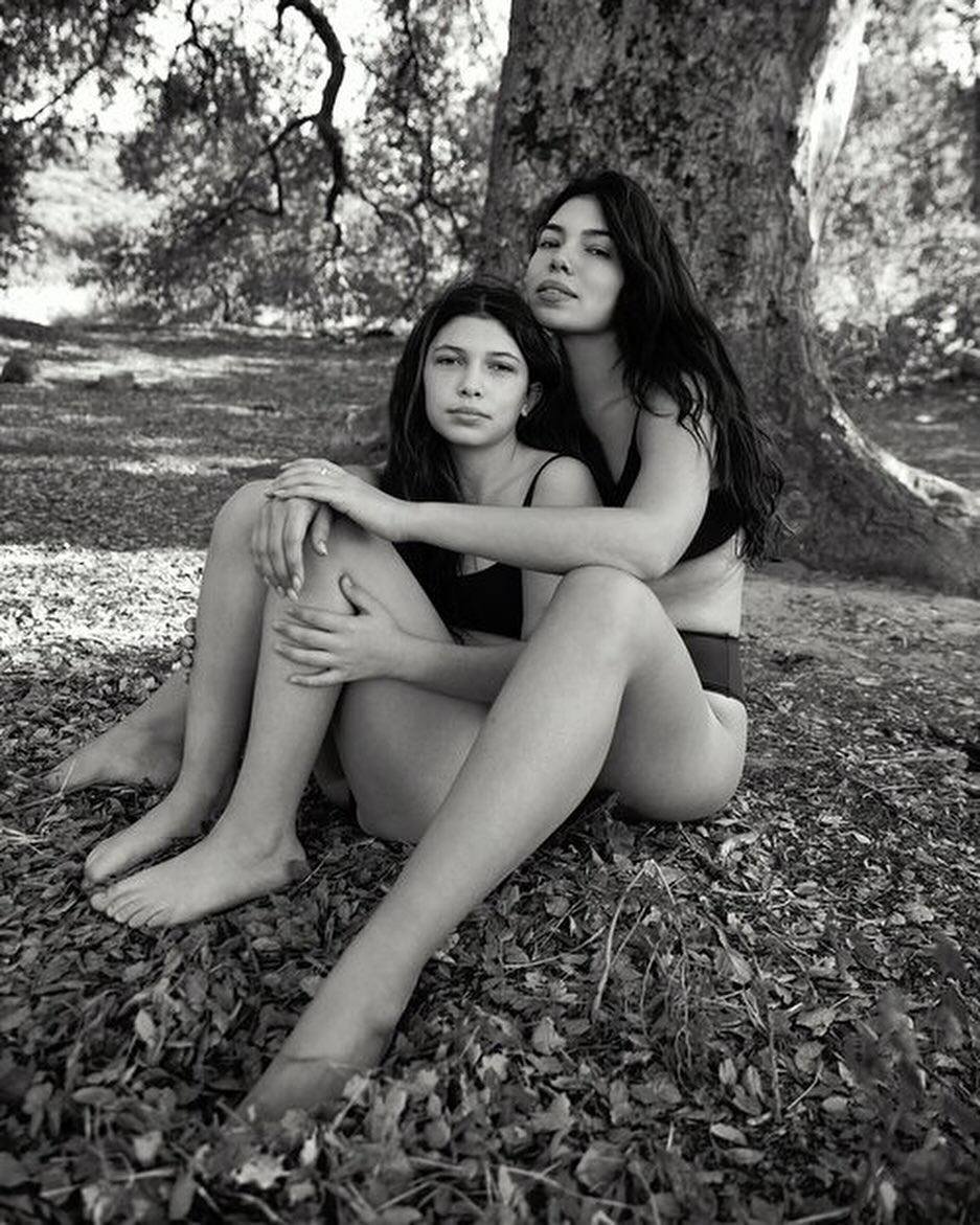 Two women seated together at the base of a large tree outdoors in a black and white portrait by Sylve Colless.