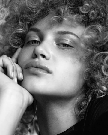 Close-up monochrome portrait of a woman with curly hair resting her chin on her hand, photographed by Sylve Colless.