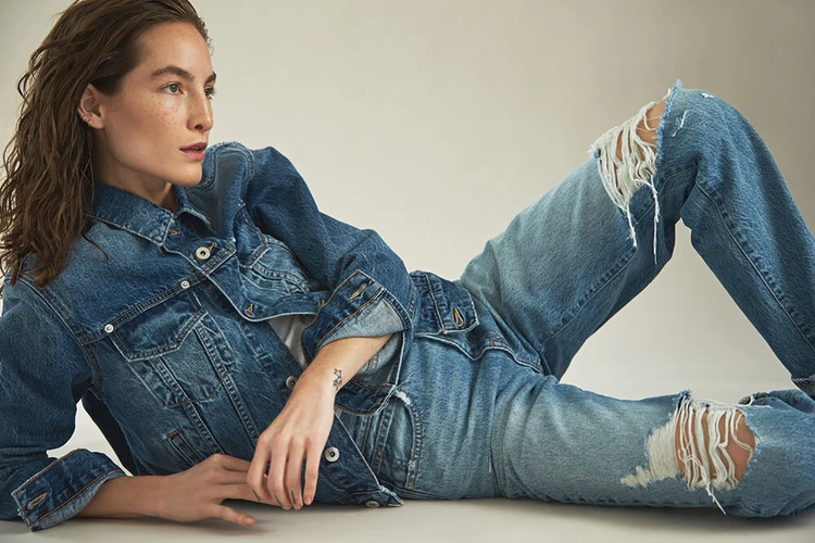 Woman in denim jacket and ripped jeans lounges in studio. Casual fashion shot with neutral background, warm lighting, and relaxed pose.