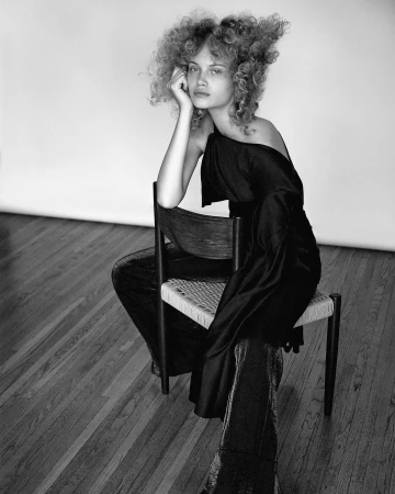 Monochrome portrait of a woman with curly hair seated backward on a wooden chair, wearing a black off-shoulder top.