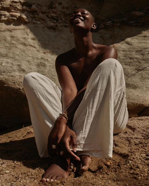 Woman sits on sandy ground, wearing light pants and smiling. Sun casts shadows on sand and rock formation backdrop.