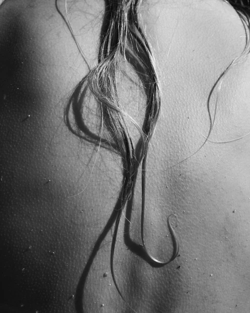 Monochrome shot of the upper back of an adult female with strands of wet hair, casting shadows, on a light outdoor background.