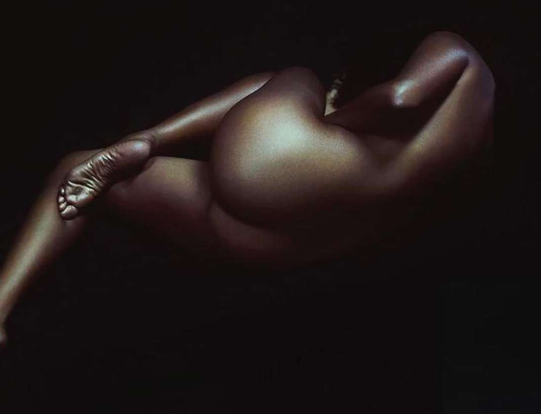 Portrait of a reclining, nude adult female with brown skin against a dark background, illuminated by studio lighting.