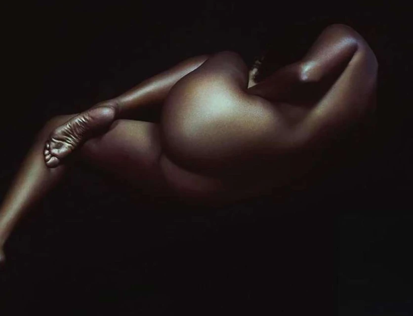 Portrait of a reclining, nude adult female with brown skin against a dark background, illuminated by studio lighting.