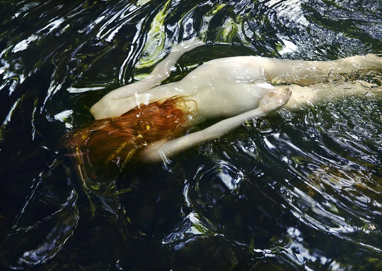 Woman with long red hair partially submerged in dark, swirling water, creating a sense of mystery and ethereal beauty in nature.