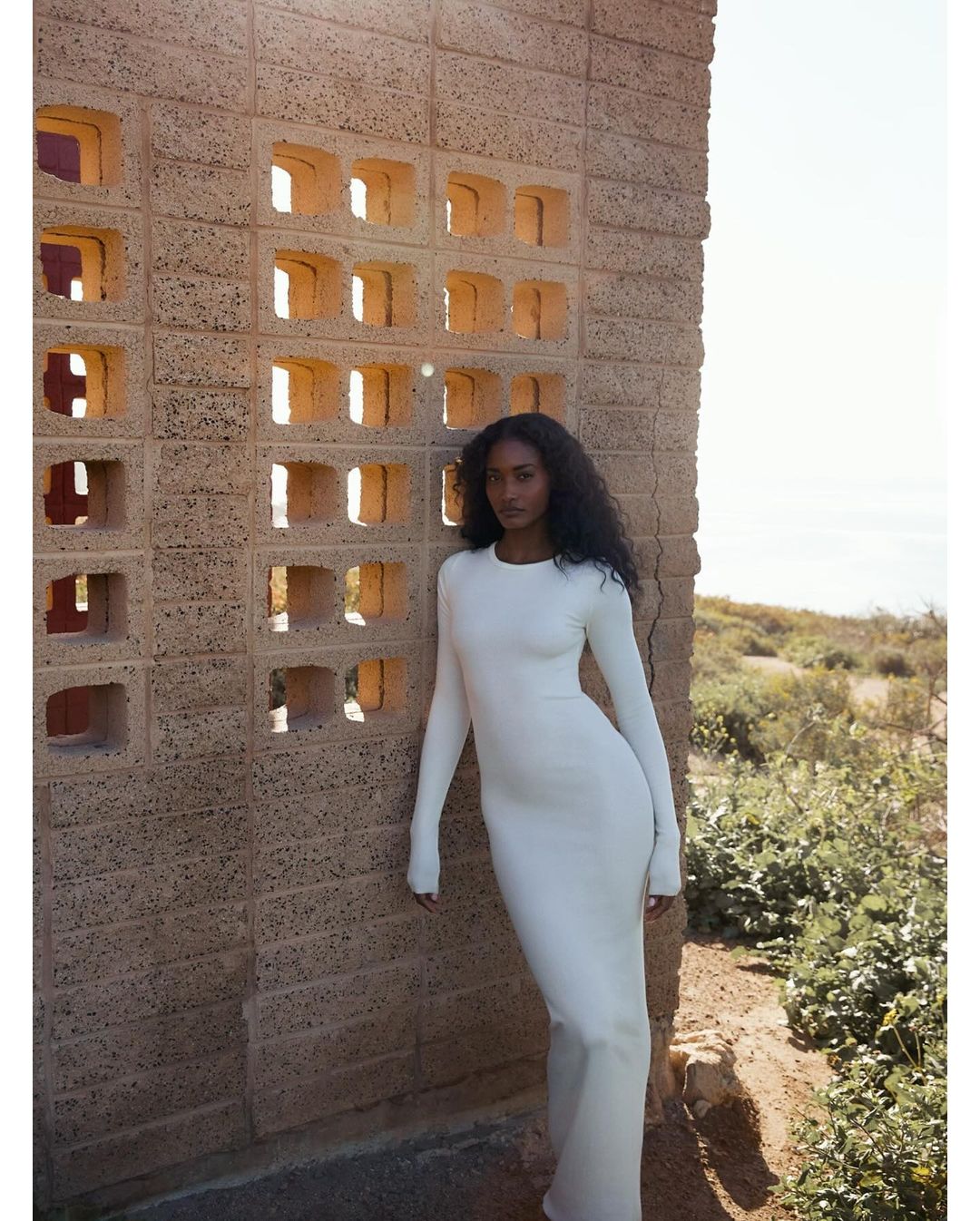 Brunette woman in long white dress poses against a unique tan brick wall on an outdoor set with ocean views in the distance.