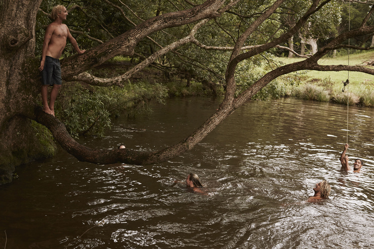 Group of people swim in a river, with one man standing on a tree branch and another swinging from a rope, all amidst a lush, green forest backdrop.