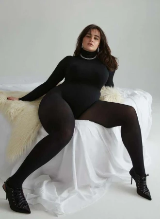 Woman in black long-sleeve bodysuit and tights seated on white bedding by Sylve Colless