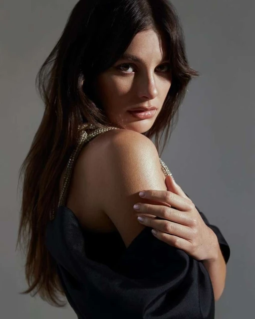 Headshot of a dark-haired woman in a black dress with bejeweled straps, looking over her shoulder in a softly lit indoor space.