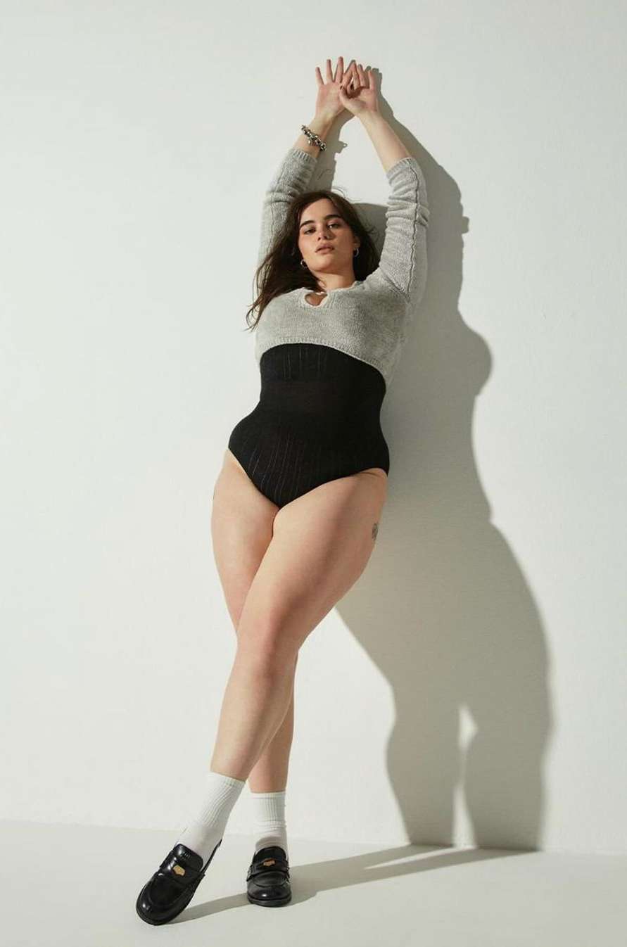 Model leaning against white wall wearing a grey top, black high waisted briefs, white socks and black loafers