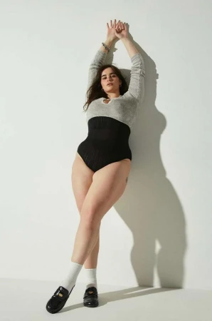 Model leaning against white wall wearing a grey top, black high waisted briefs, white socks and black loafers