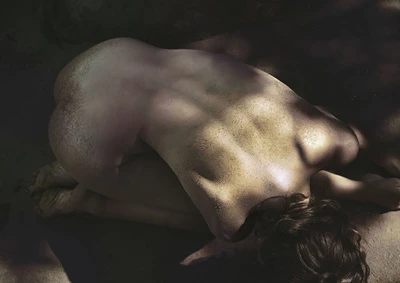 A person curled up on a sandy surface with dappled shadows across their back, captured from a high angle by photographer Sylve Colless.