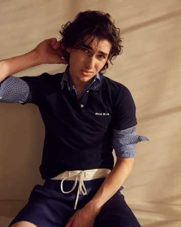 Adult male seated portrait: man with dark curly hair poses in a navy blue polo shirt and blue shorts in a softly lit photo studio.