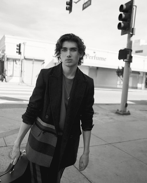 Black and white portrait of a young man in a jacket and carrying a case on a city street with traffic lights and buildings.