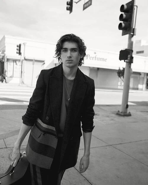 Black and white portrait of a young man in a jacket and carrying a case on a city street with traffic lights and buildings.