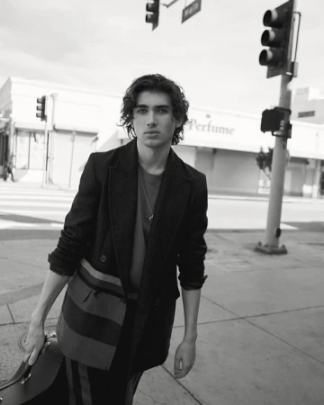 Black and white portrait of a young man in a jacket and carrying a case on a city street with traffic lights and buildings.