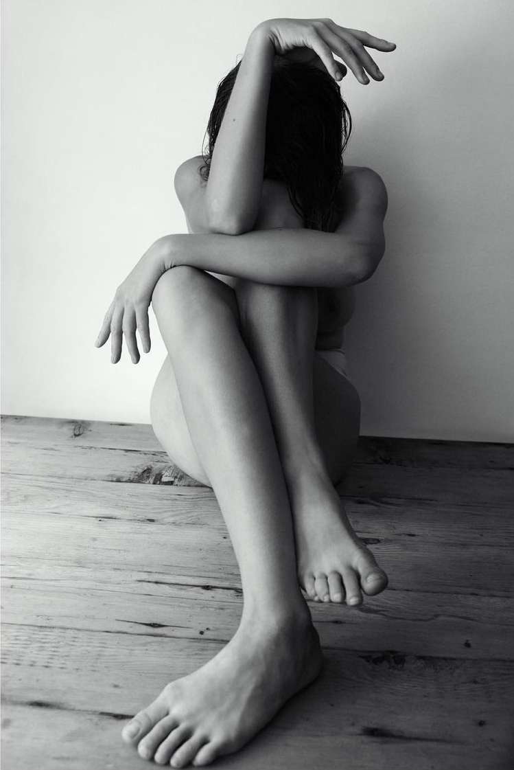 Black and white portrait of a seated woman with obscured face, legs extended, resting against a wall on a weathered wooden floor.