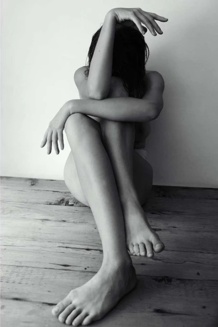 Black and white portrait of a seated woman with obscured face, legs extended, resting against a wall on a weathered wooden floor.