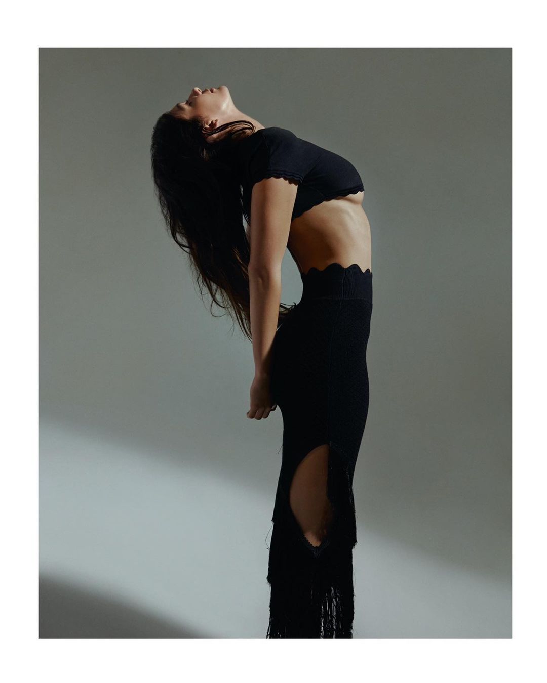 Adult woman with dark hair in a black two-piece dress arched back, posing in a studio with diffused lighting and a grey background.