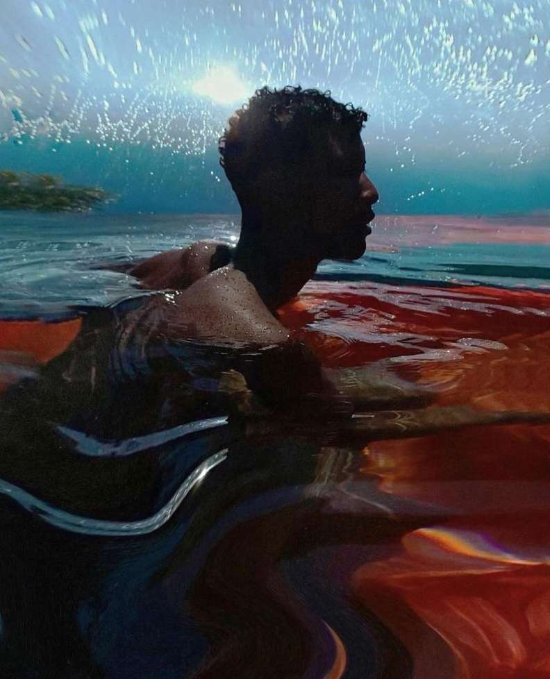 Adult man in water with orange fabric, ocean backdrop. Side view portrait with dark hair, ripples, and splashes in an aquatic scene.