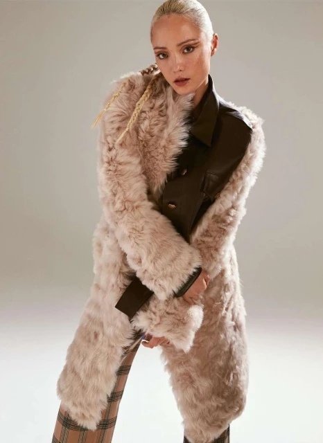 Young woman poses wearing a long, tan faux fur coat with a dark brown leather collar and cuffs, plus plaid pants against a grey backdrop.