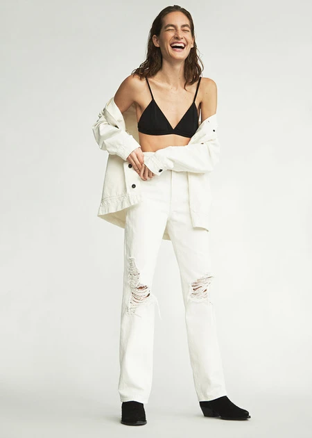 A carefree woman with a joyful laugh is wearing an unbuttoned white denim jacket and distressed white denim jeans against a white background.
