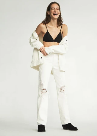 A carefree woman with a joyful laugh is wearing an unbuttoned white denim jacket and distressed white denim jeans against a white background.