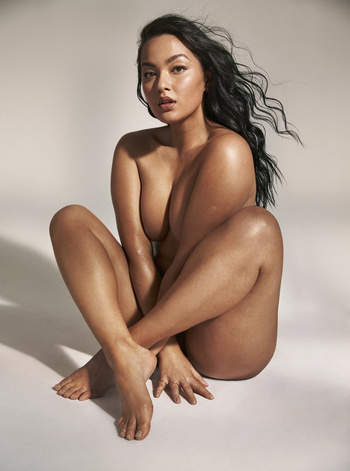 An adult female poses in a photo studio. She is nude and seated with her legs crossed and arms resting on her legs. Her dark hair is windswept.