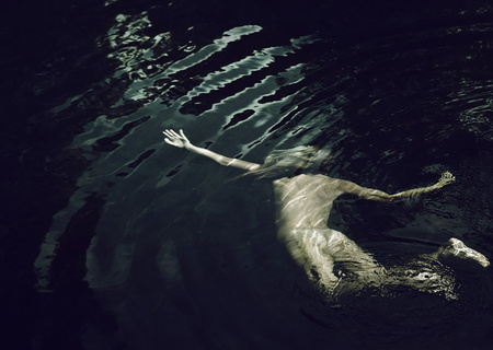 Overhead view of a person swimming in dark water with rippling caustic light patterns across their submerged body.
