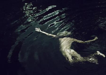 Overhead view of a person swimming in dark water with rippling caustic light patterns across their submerged body.