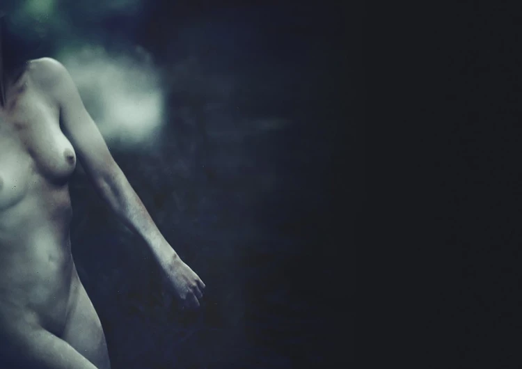 An adult woman with bare skin stands partially visible against a dark and smoky backdrop in a studio photography setting.