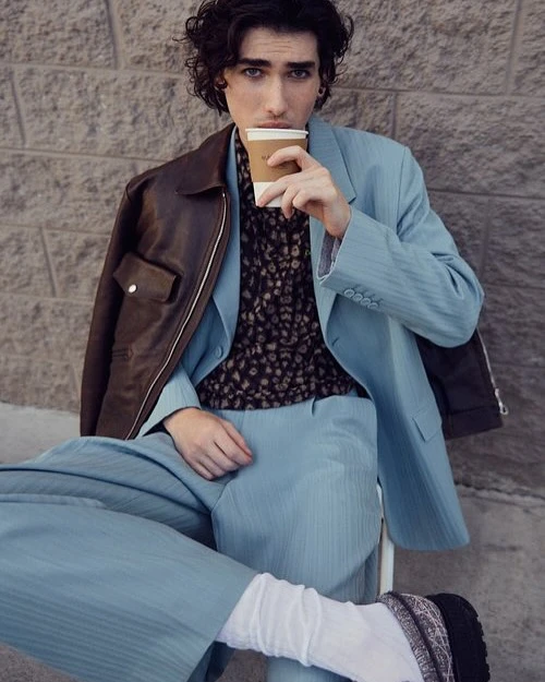 Young man in a light blue suit sits against a brick wall while holding a coffee cup, wearing a patterned shirt and leather jacket.