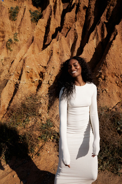 Woman in long sleeve dress poses outdoors against eroded earth. Smiling woman in white dress with geological background on a sunny day.