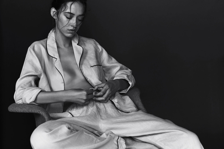 Monochromatic portrait of a young adult woman in pajama-style clothing, seated in a chair against a dark background, looking down.