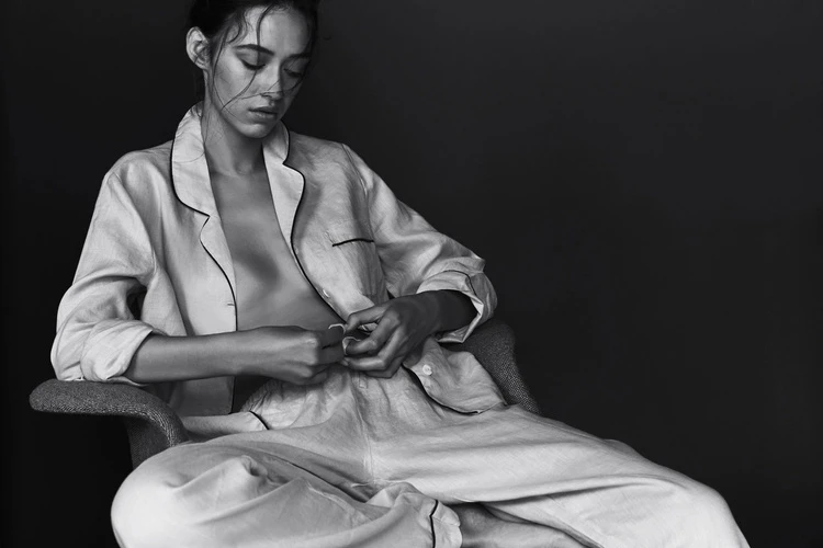 Monochromatic portrait of a young adult woman in pajama-style clothing, seated in a chair against a dark background, looking down.