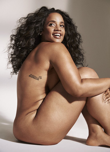 Portrait of a smiling nude adult female with curly hair sitting with her arms around her legs in a photo studio with a light beige backdrop.