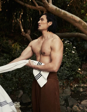 Shirtless Asian male poses with a towel in a natural outdoor setting surrounded by trees and foliage, wearing a brown wrap skirt.